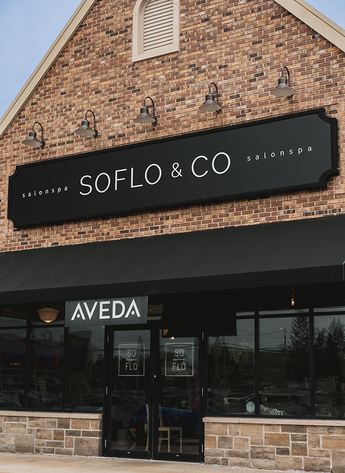 Front angled view of SoFlo & Co North location in Moncton, exterior. Bricks and black sign as well as an AVEDA logo over the doors.